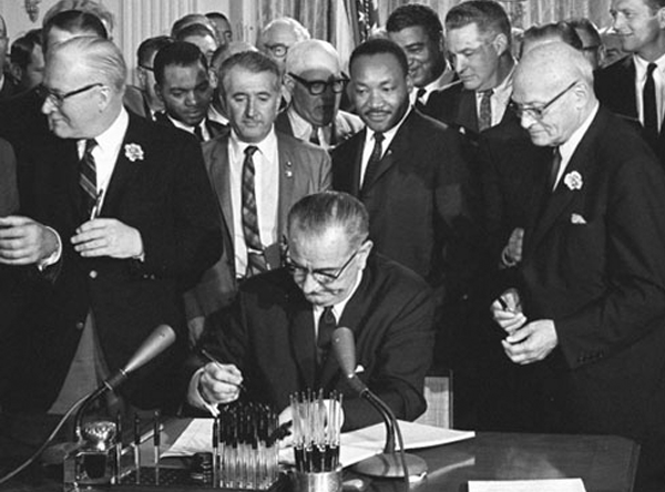 Lyndon B. Johnson signs the Civil Rights Act of 1964. Martin Luther King, Jr. stands behind the President.